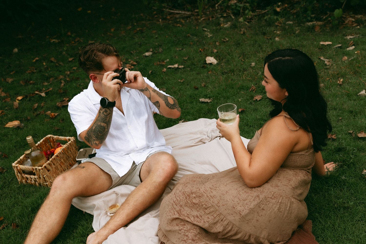 Couple enjoying a picnic on grass, man with tattoos photographing woman holding a drink on a blanket.