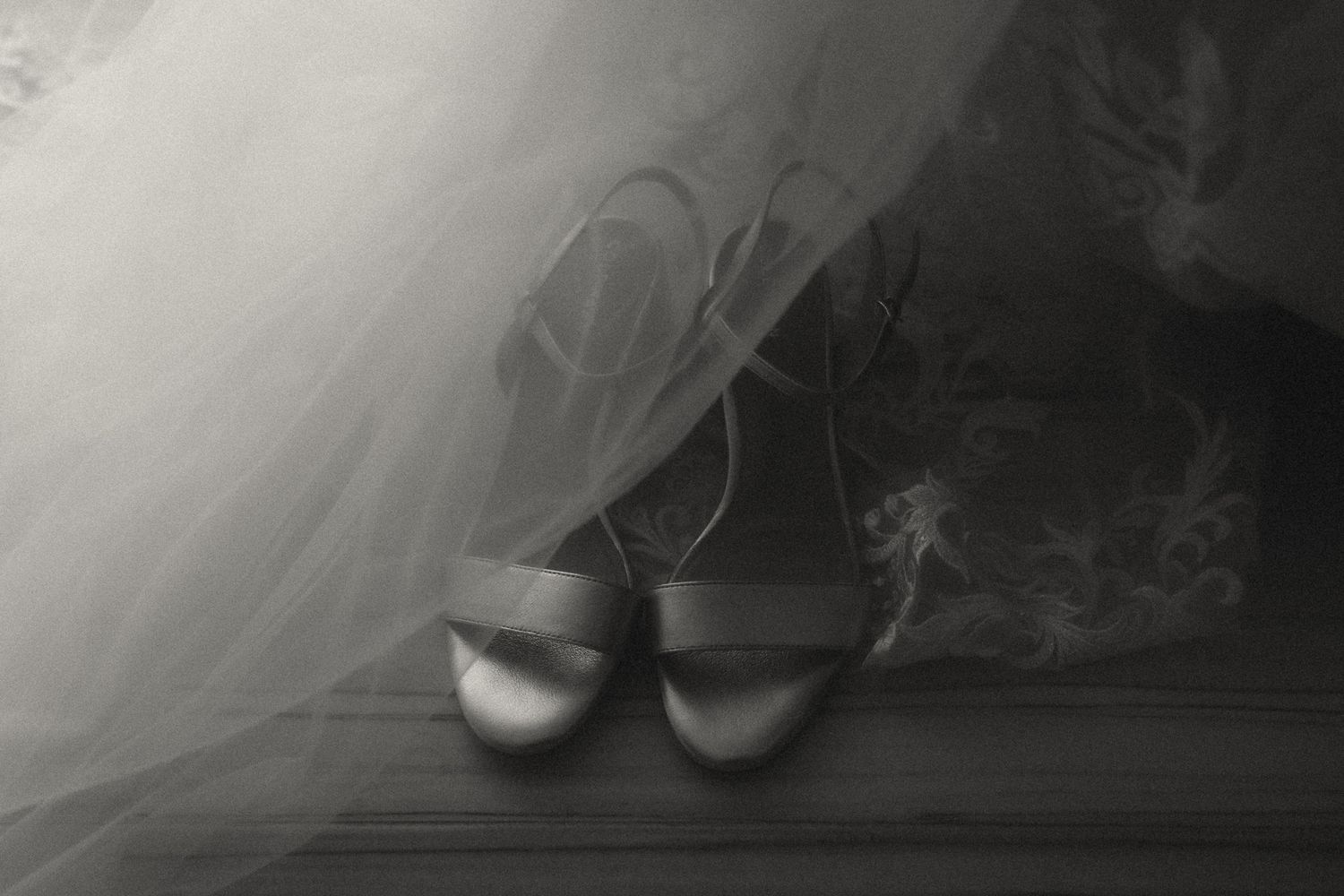 Black and white photo of satin bridal shoes peeking under a flowing wedding veil on a wooden floor.