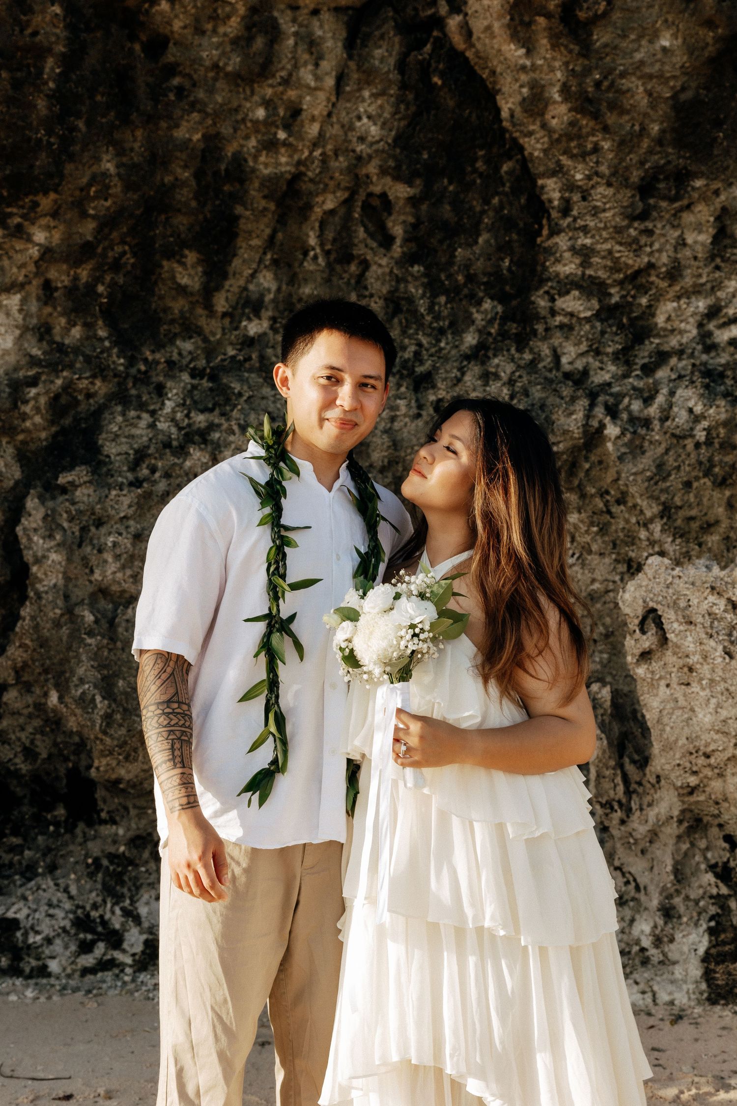 Hawaiian wedding couple in white attire, groom wearing green lei, bride holding white bouquet, posing before rocky cliff backdrop.