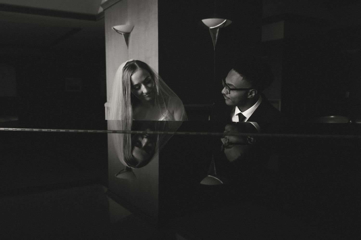 Black and white photo of bride and groom sharing an intimate moment at a piano, reflections visible on polished surface.