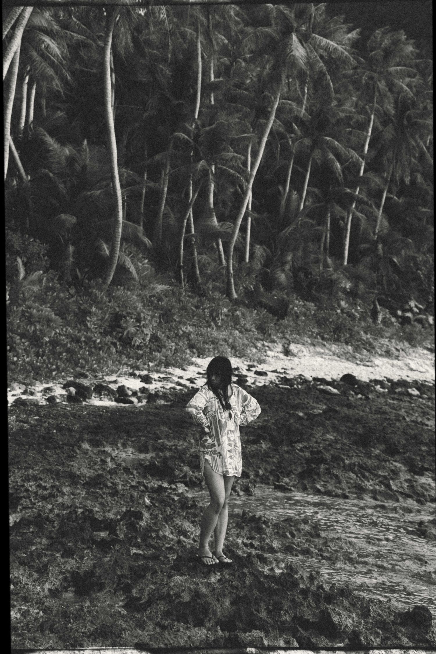 Black and white photo of a woman standing on a rocky tropical beach surrounded by tall palm trees.