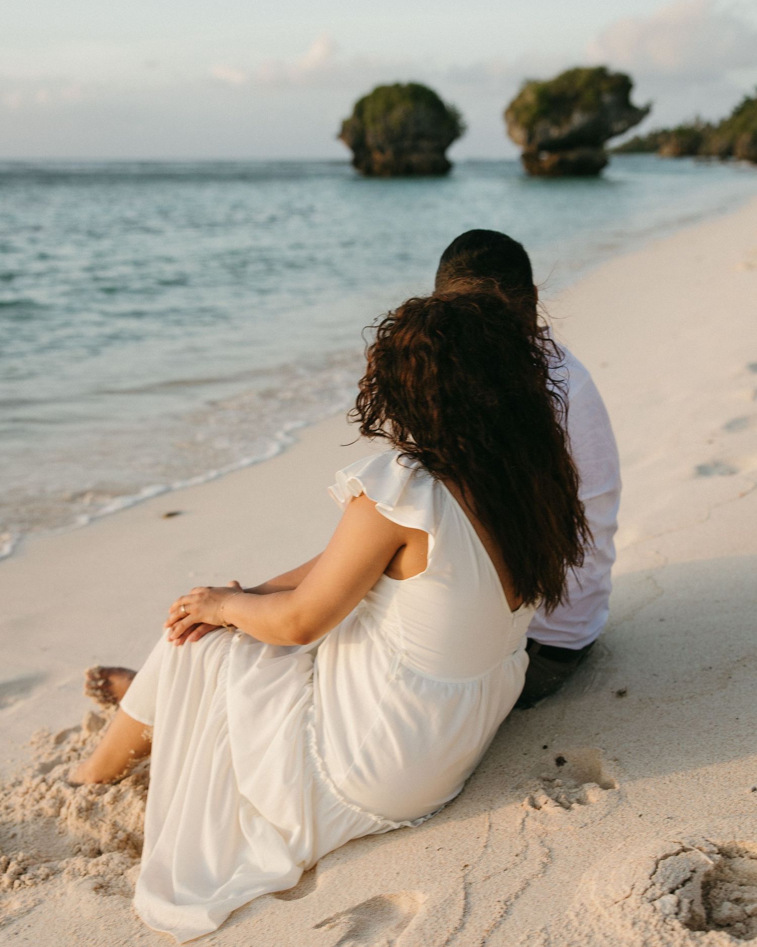 Couple sitting together on a white sandy beach, gazing at rocky ocean formations in the distance at sunset.
