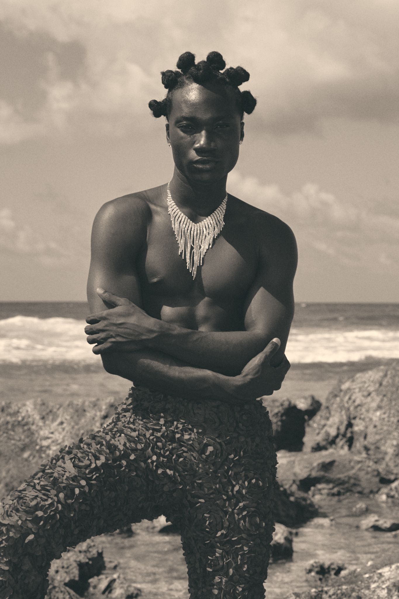 Shirtless muscular man with bun hairstyle and beaded necklace posing on rocky beach in black and white photo.