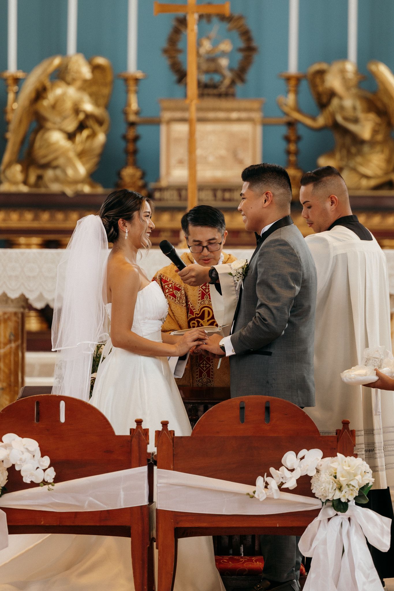 Couple exchanging rings at Catholic church altar during wedding ceremony with officiant and golden angel decor.