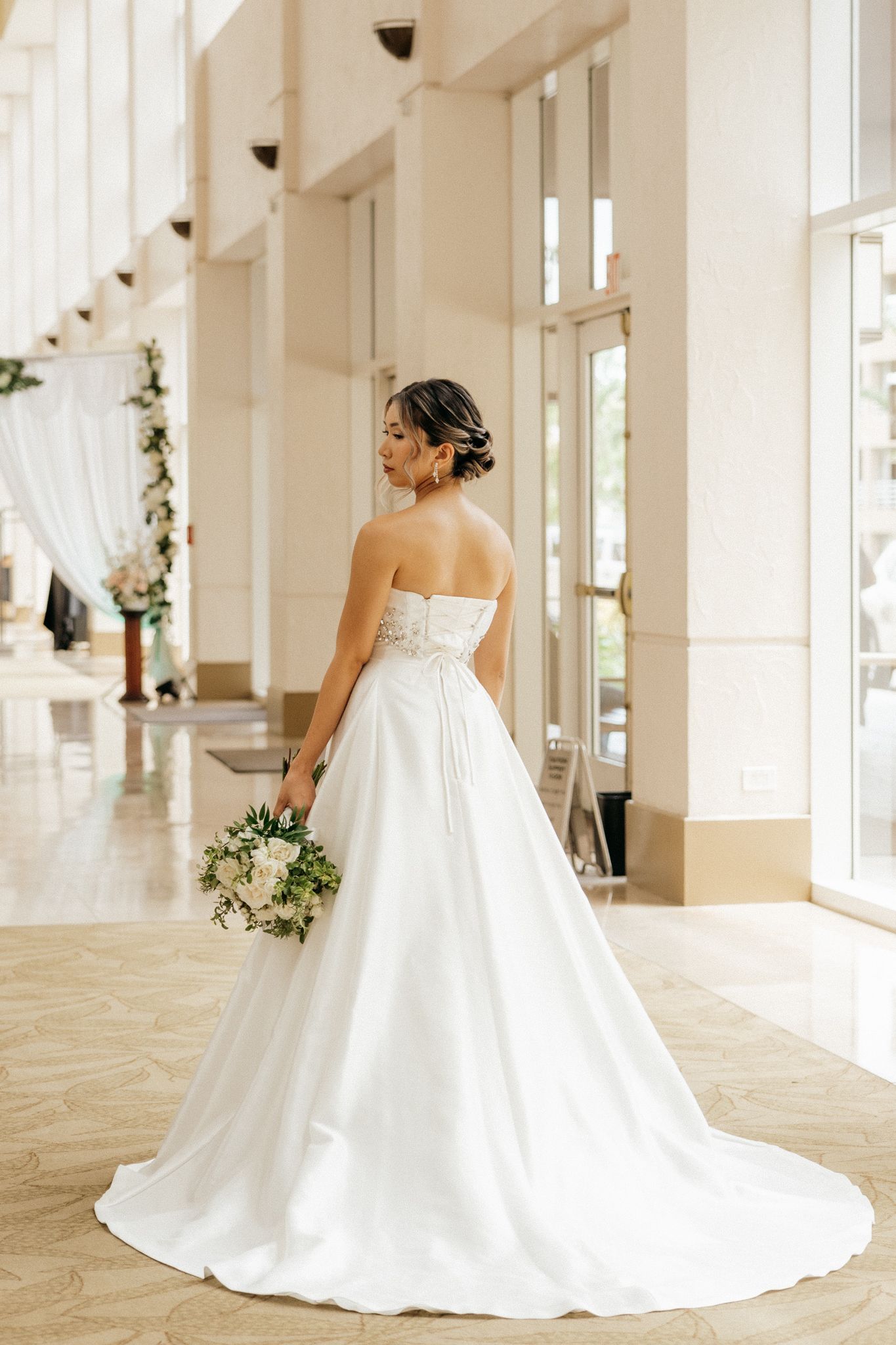 Bride in strapless white ball gown with train holds bouquet, viewed from behind in elegant wedding venue.