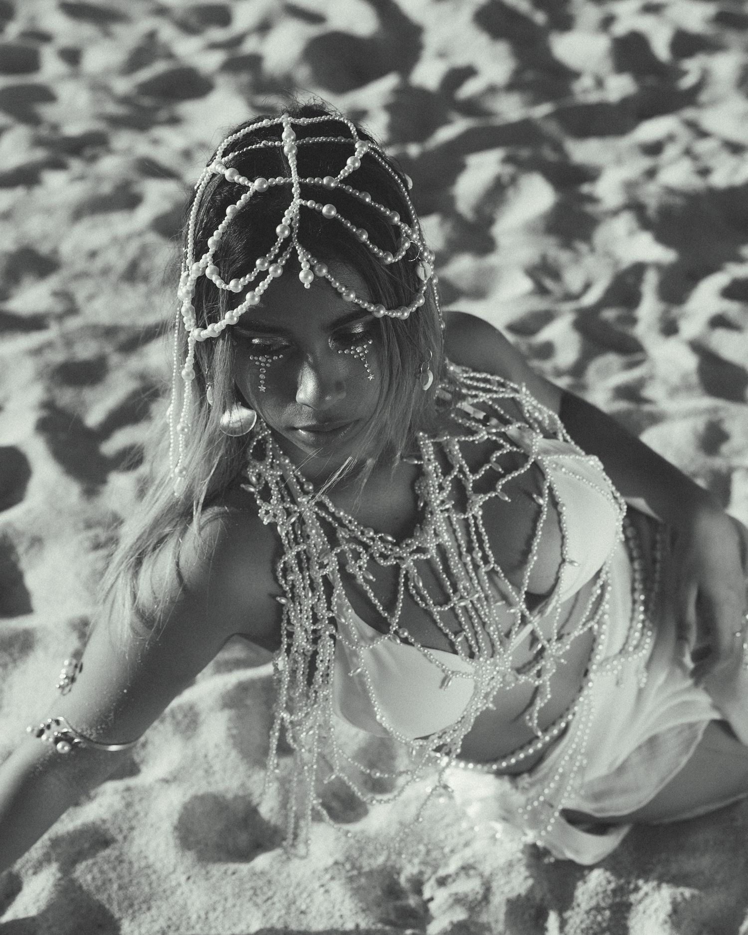 Woman adorned with pearl headpiece and jewelry poses on sandy beach in black and white photo.
