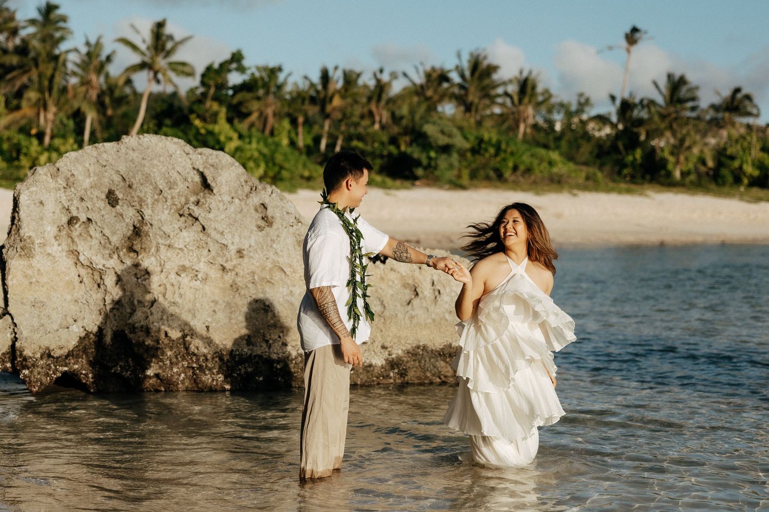 Couple in wedding attire wading in tropical beach water, bride in white dress, groom in linen suit with floral lei.
