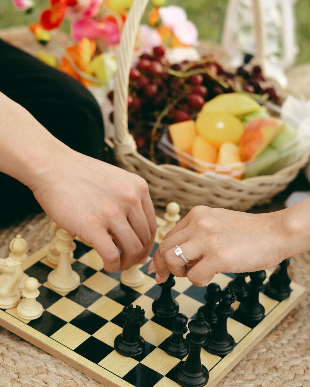 Two people playing chess at a picnic with a fruit basket in the background.