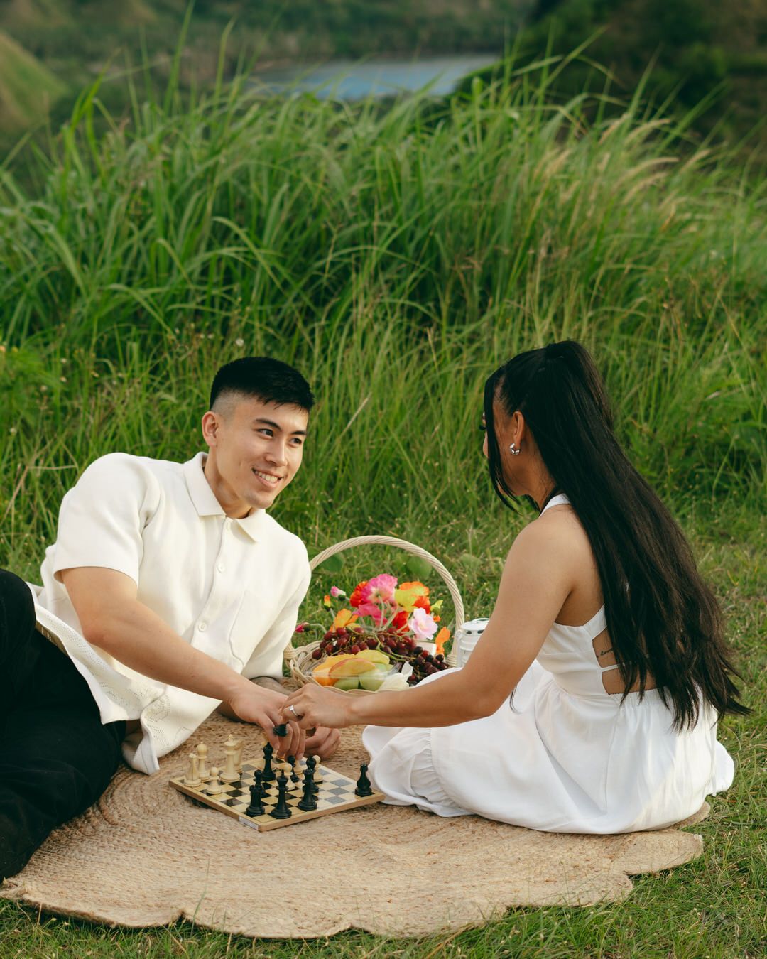 Couple enjoying a romantic outdoor picnic on a blanket with a flower basket and chess board surrounded by lush green grass.