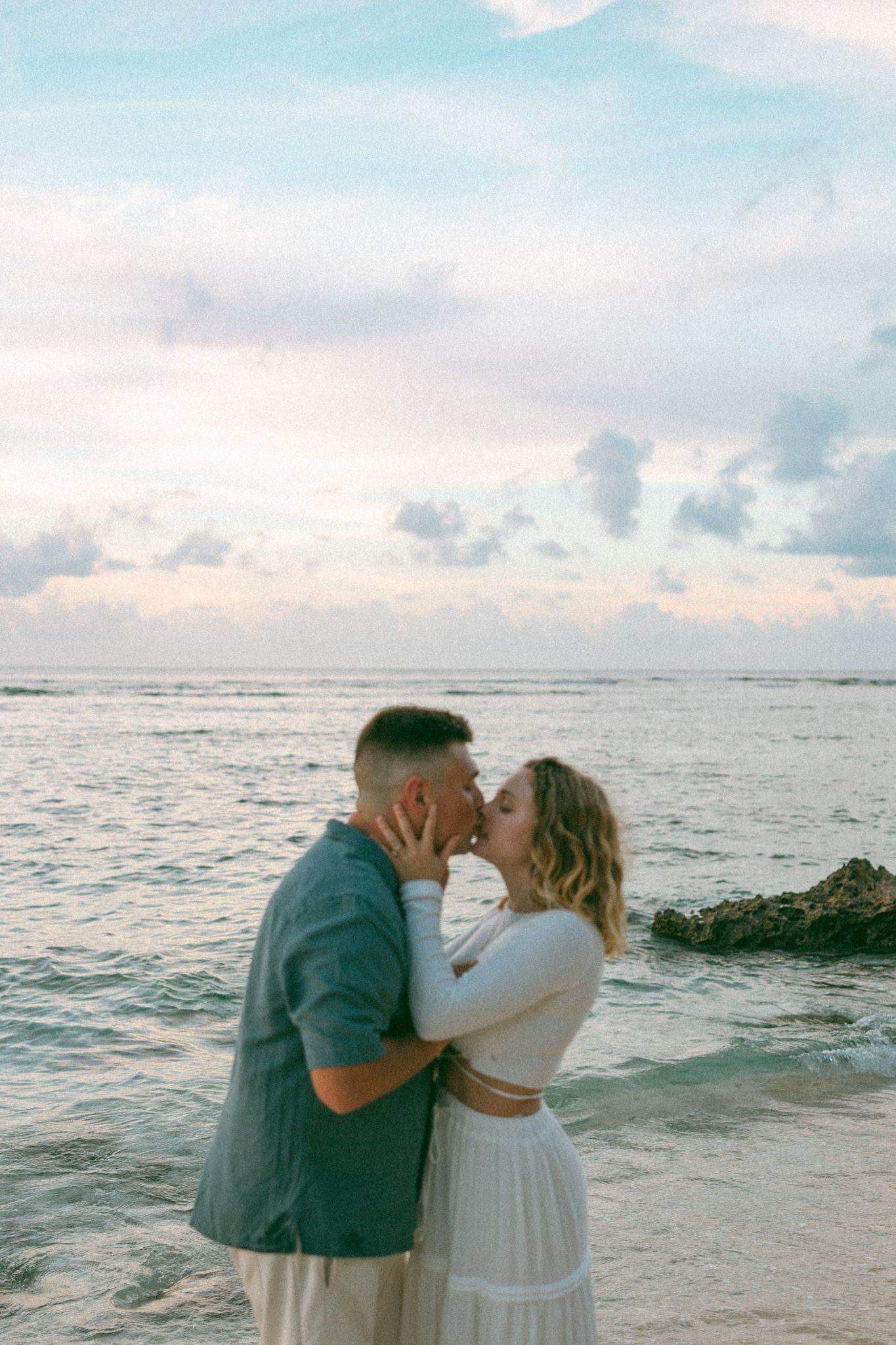Couple sharing a romantic kiss on a tropical beach at sunset with pastel sky and ocean waves.
