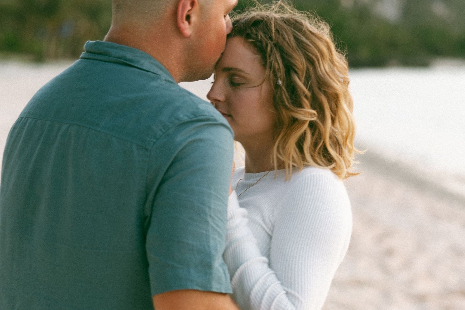 Romantic couple sharing an intimate moment on a sandy beach, with the woman in a white top embracing her partner.