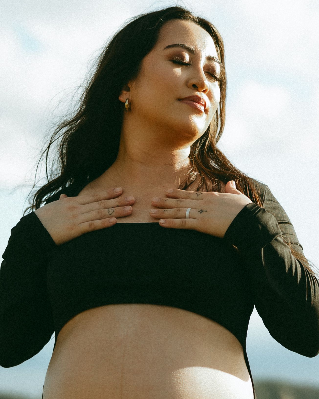 Pregnant woman in black crop top holding hands to chest outdoors with cloudy sky background.