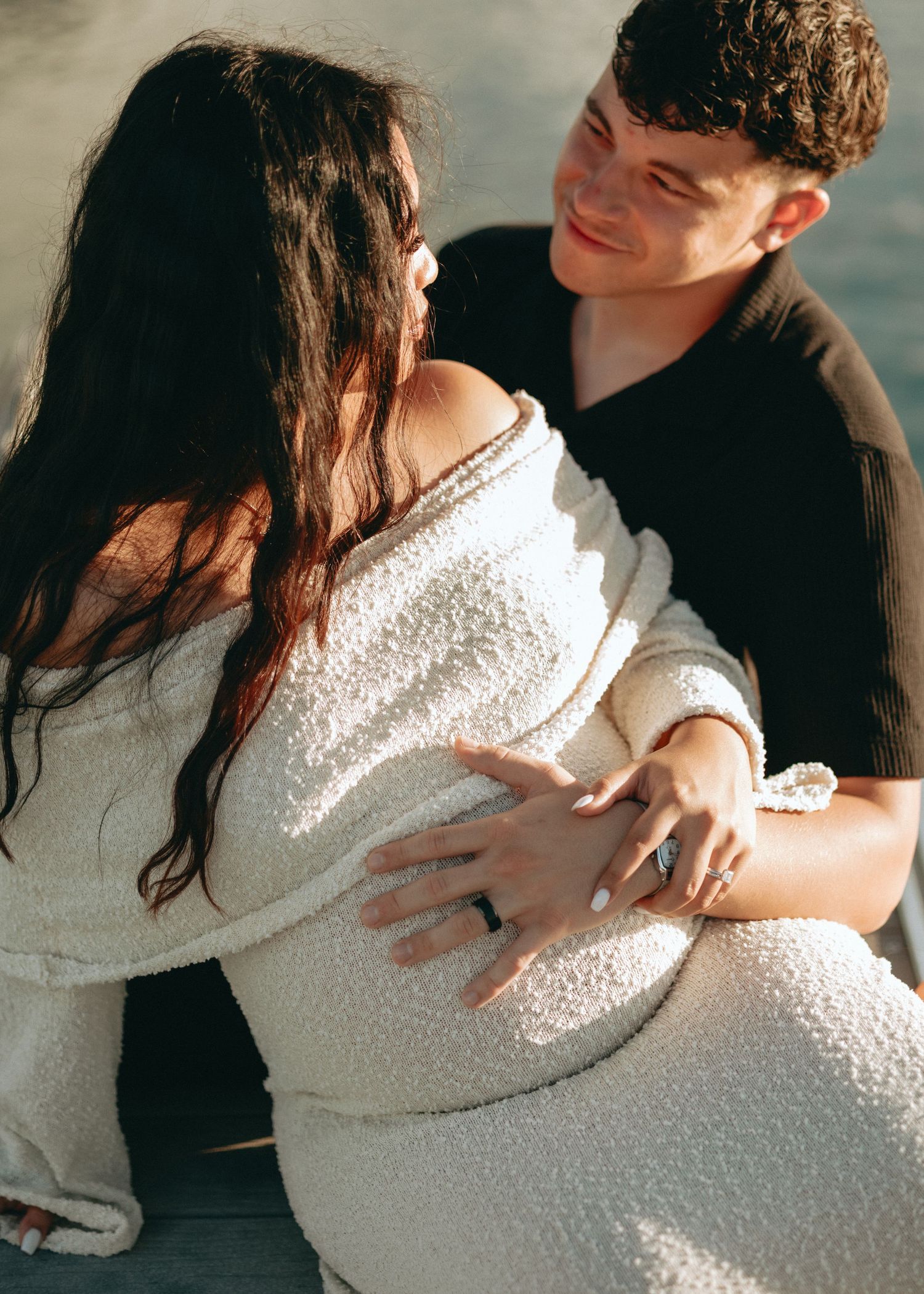 Couple sharing a tender moment, woman in white boucle off-shoulder dress, man in black shirt, sitting closely together outdoors.