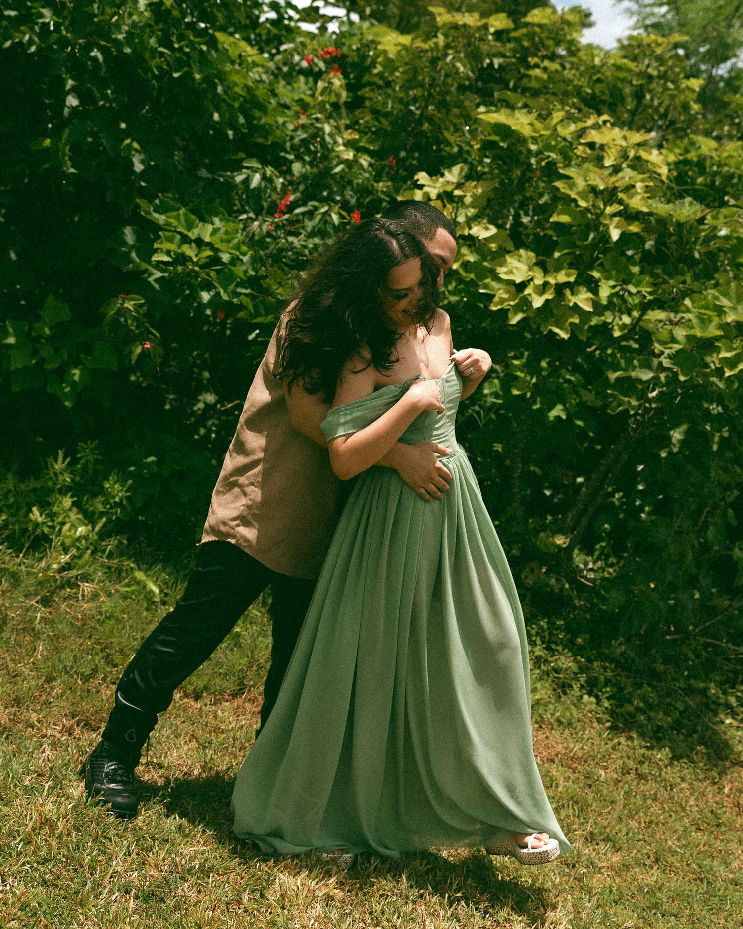Couple embracing outdoors, woman in sage green gown, man in tan jacket, surrounded by lush green foliage.