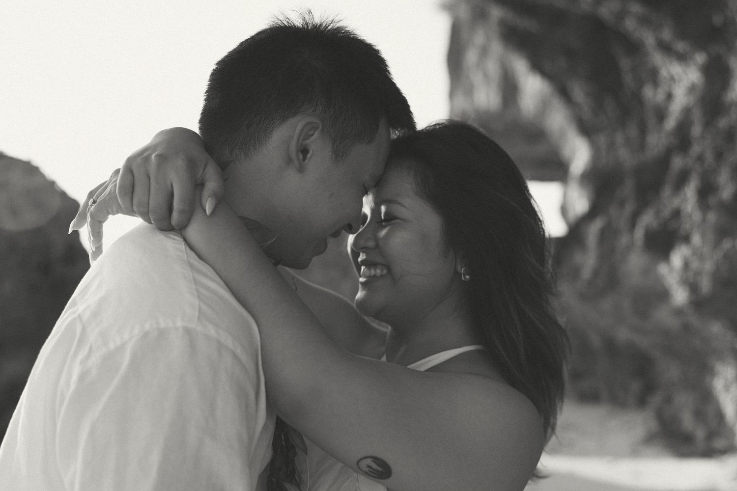 Black and white photo of a romantic couple sharing an intimate moment on a beach near rocky cliffs.