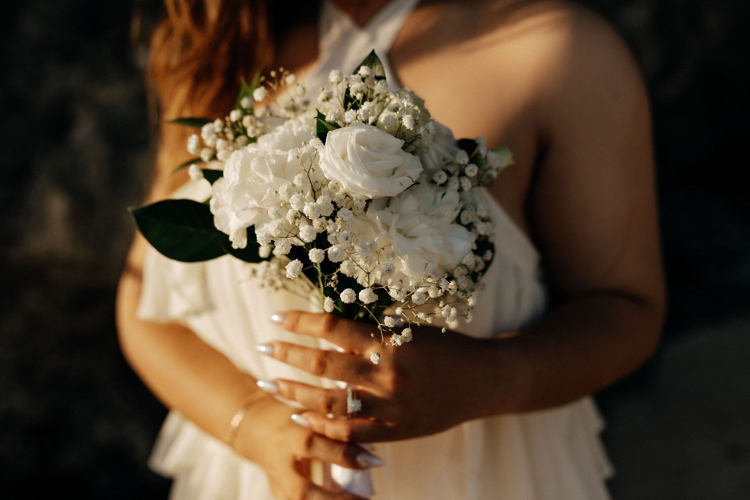 Bride holding a white wedding bouquet of roses and baby's breath, wearing a ring on her finger.