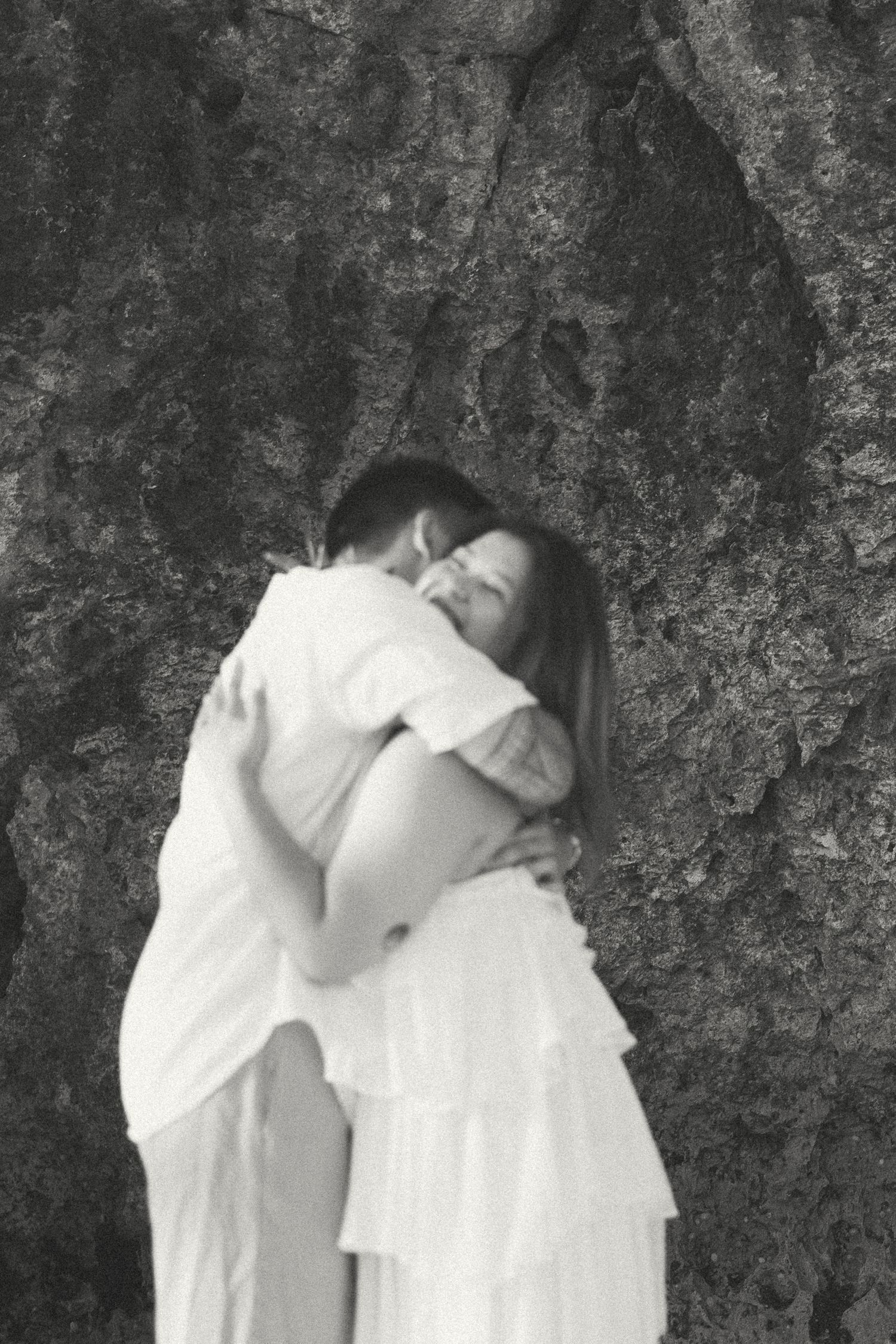 Black and white photo of a couple embracing and smiling against a textured rock wall, wearing white outfits.