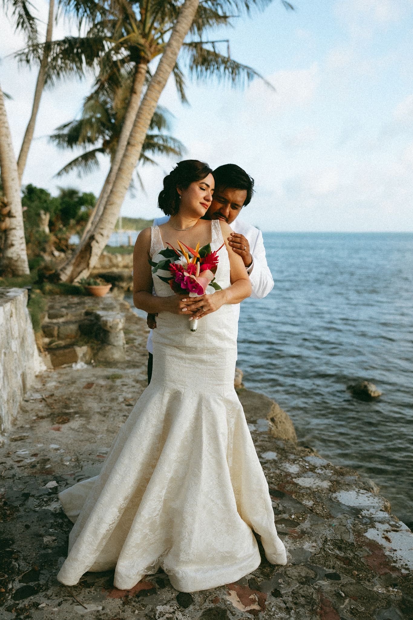 Bride and groom embrace on rocky beachside with palm trees and ocean backdrop during tropical destination wedding.