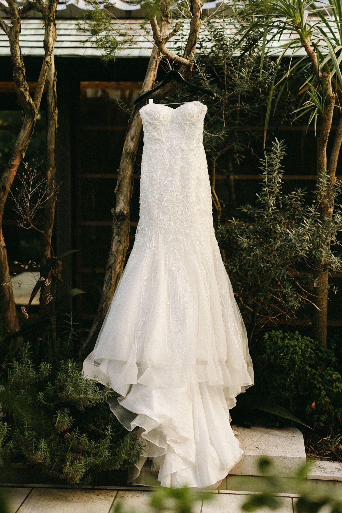 Elegant strapless mermaid wedding dress with lace bodice and layered skirt hanging outdoors among lush greenery.