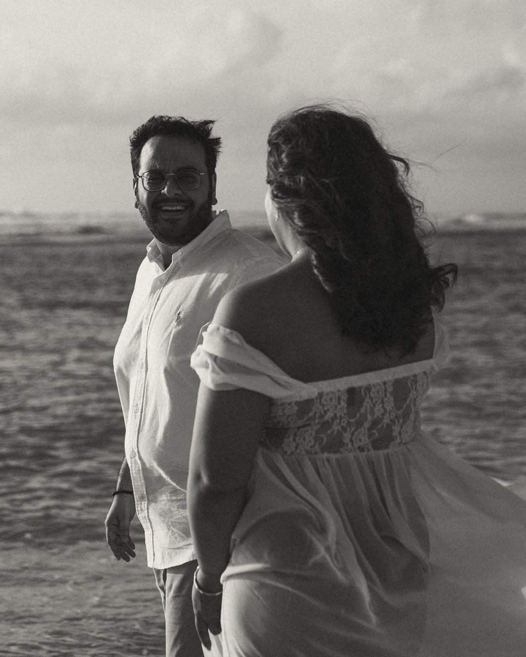 Black and white photo of a smiling man in white shirt and woman in flowing dress standing by the ocean.