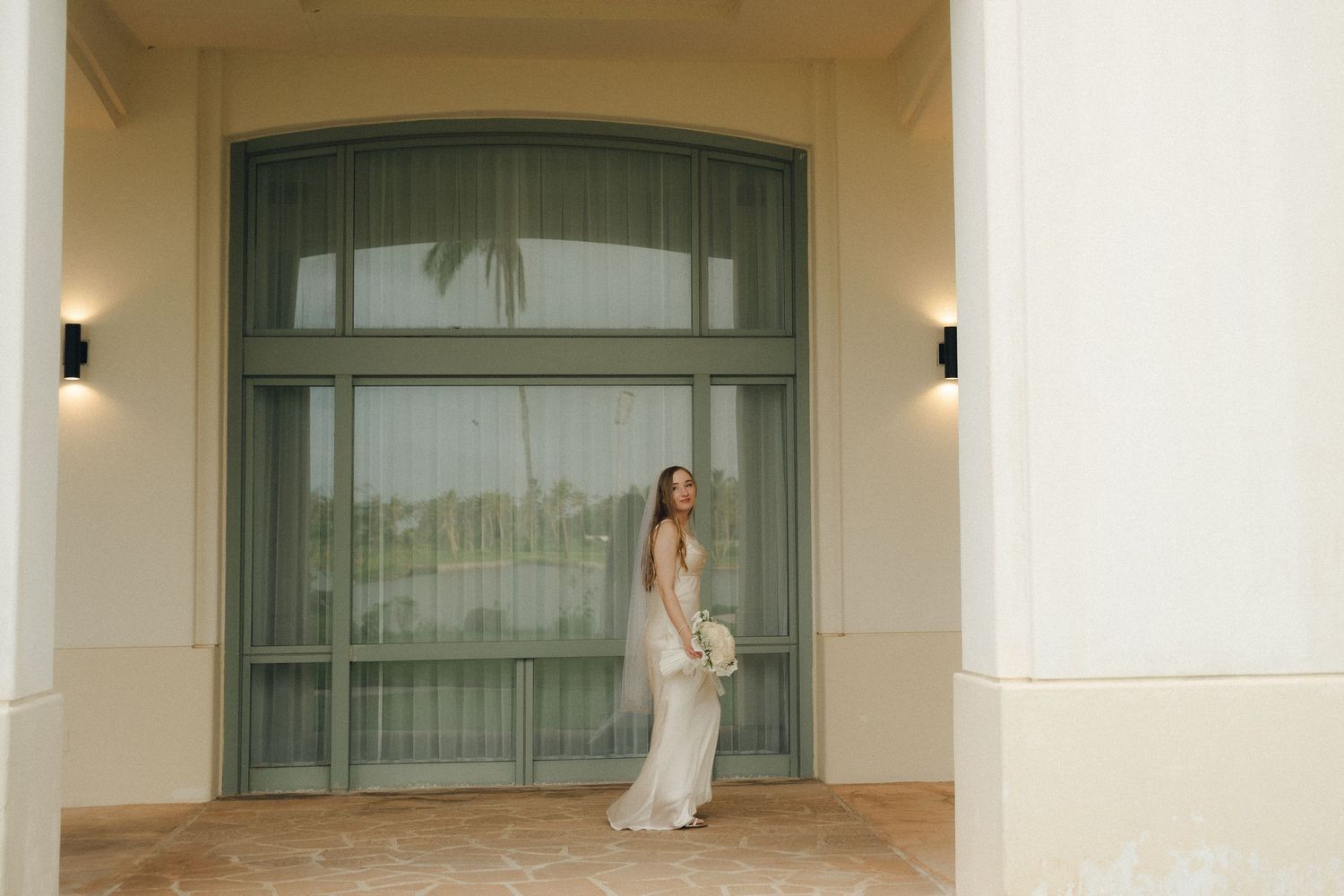 Bride in elegant white gown poses by green-framed glass doors at a luxury venue with tropical views.