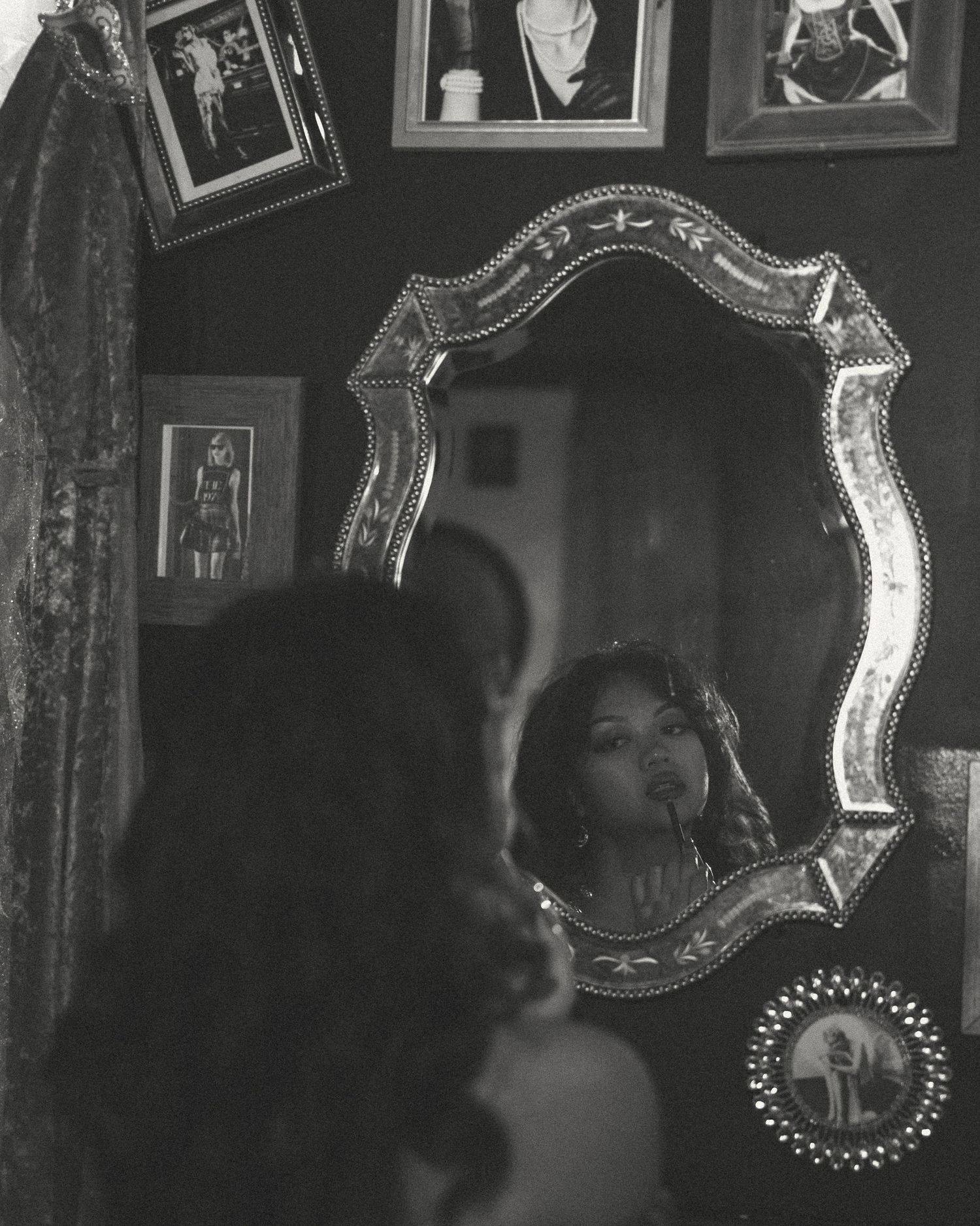 Black and white photo of a woman gazing at her reflection in an ornate vintage mirror, surrounded by framed pictures on a dark wall.