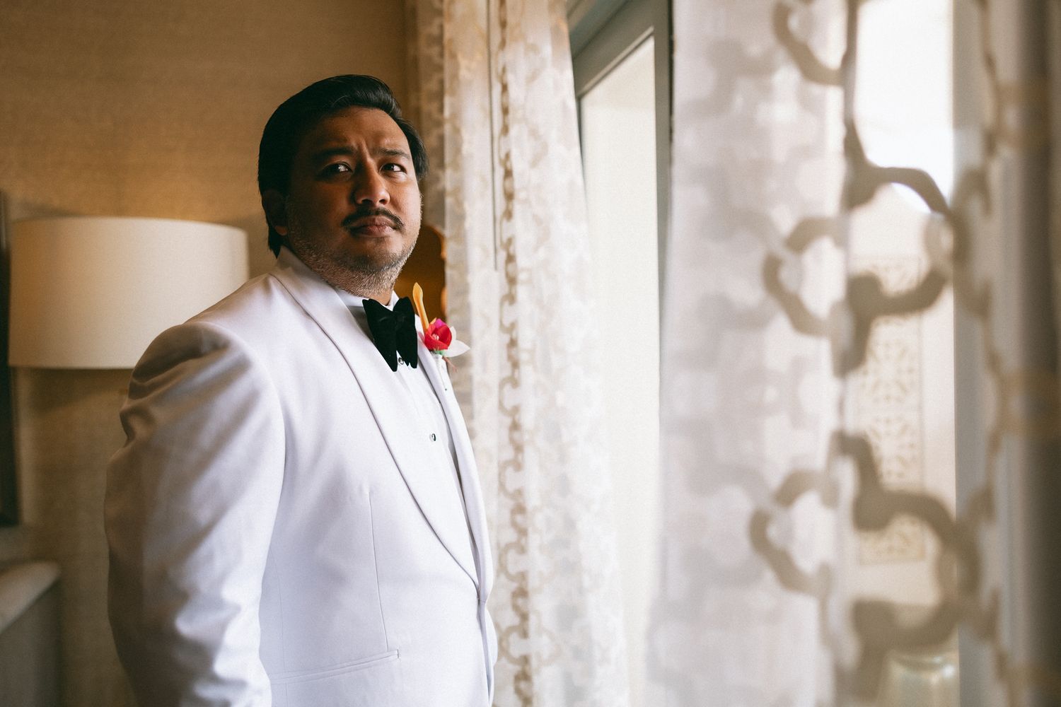 Groom in white tuxedo with red boutonniere standing by a curtained window in elegant wedding attire.