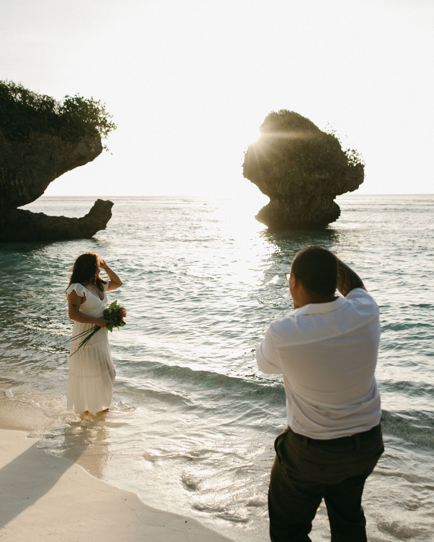Couple having wedding photos taken on a tropical beach at sunset with rocky formations in the ocean.