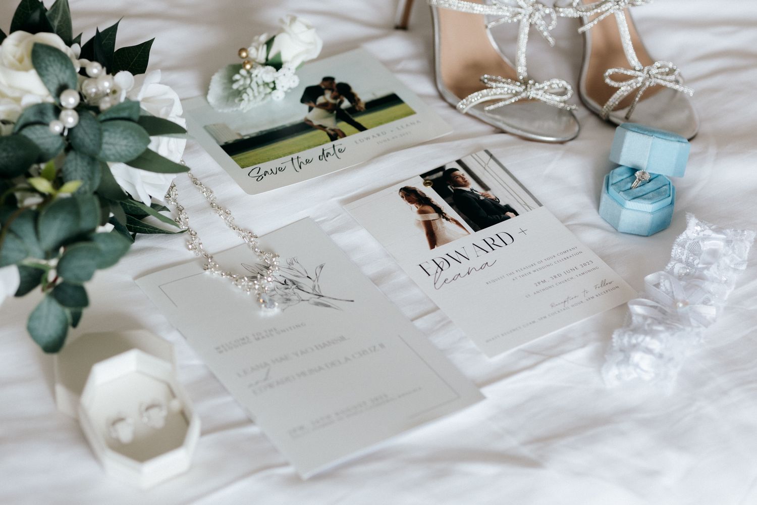 Elegant wedding flat lay featuring invitations, save the date cards, bridal shoes, floral bouquet, and accessories on white fabric.