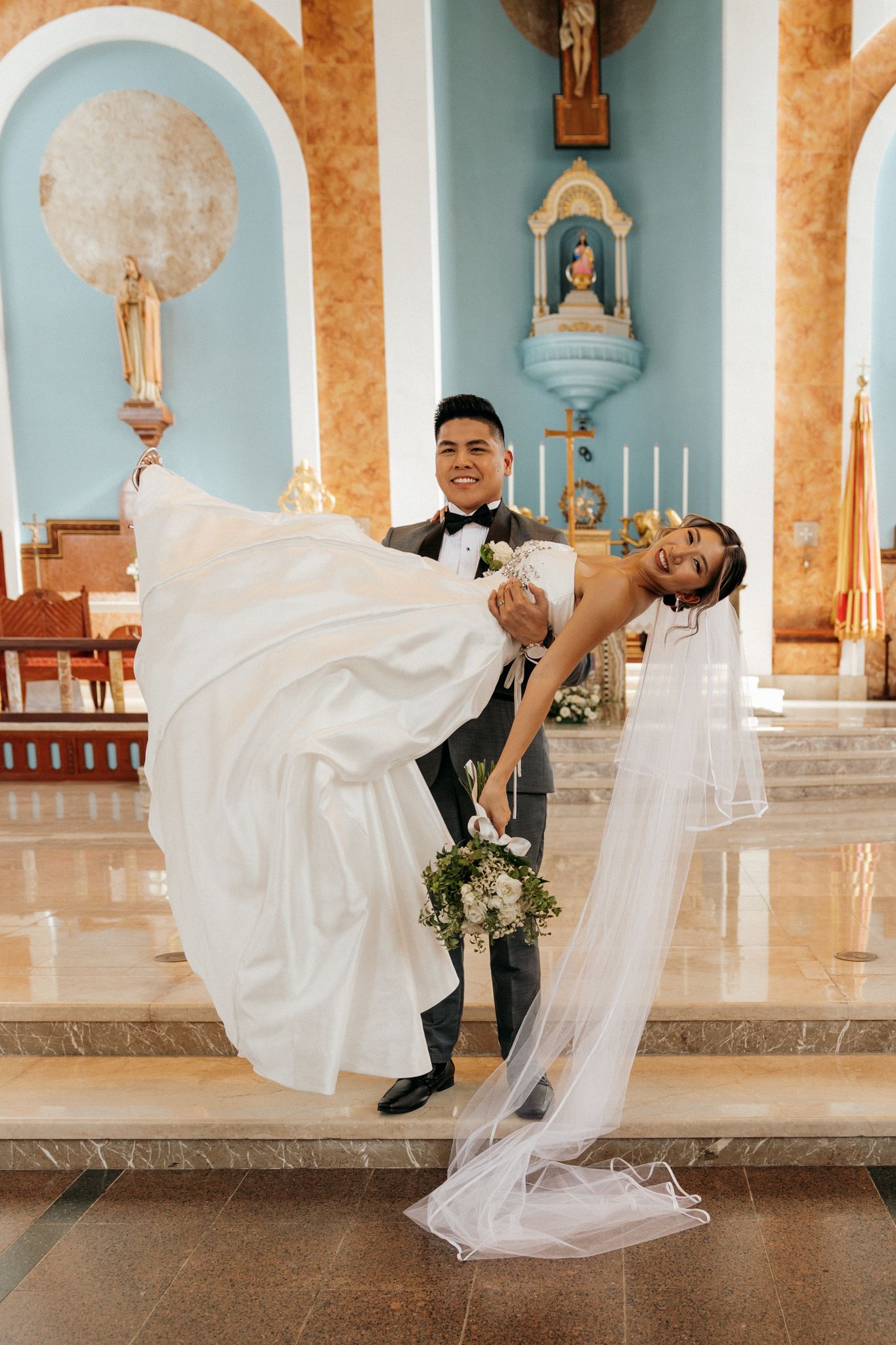 Groom lifts bride in white gown and veil at church altar, surrounded by blue walls and religious decor.
