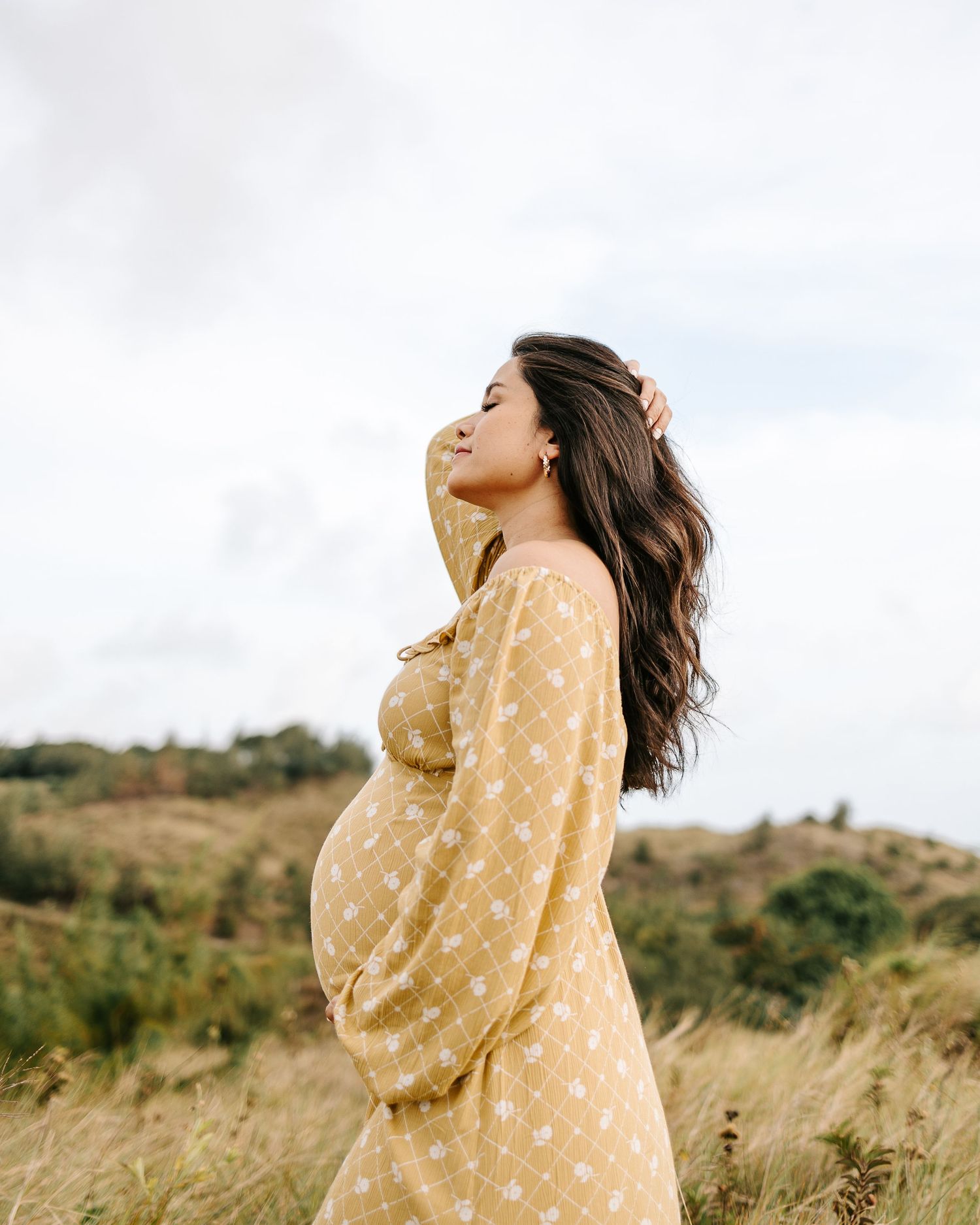 Pregnant woman in yellow floral dress standing in a field, posing sideways with hand in hair against cloudy sky.