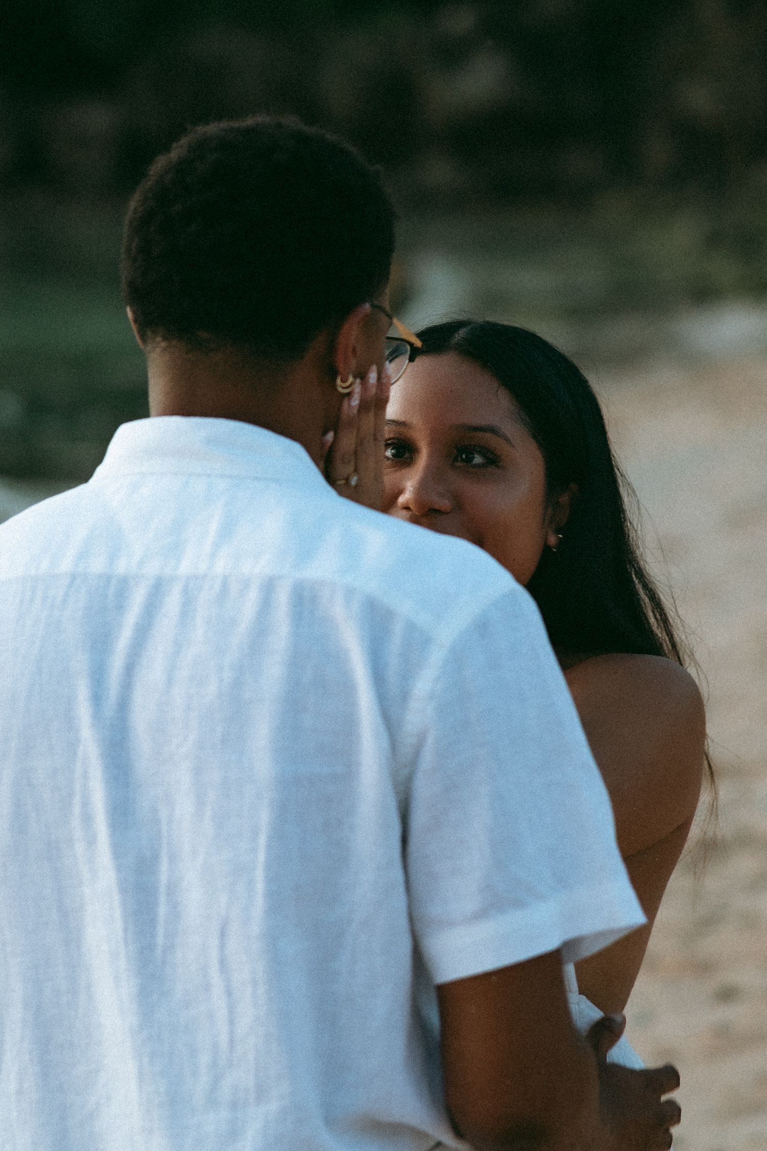 A woman peeks over a man's shoulder as they embrace on a beach, both dressed casually in warm evening light.