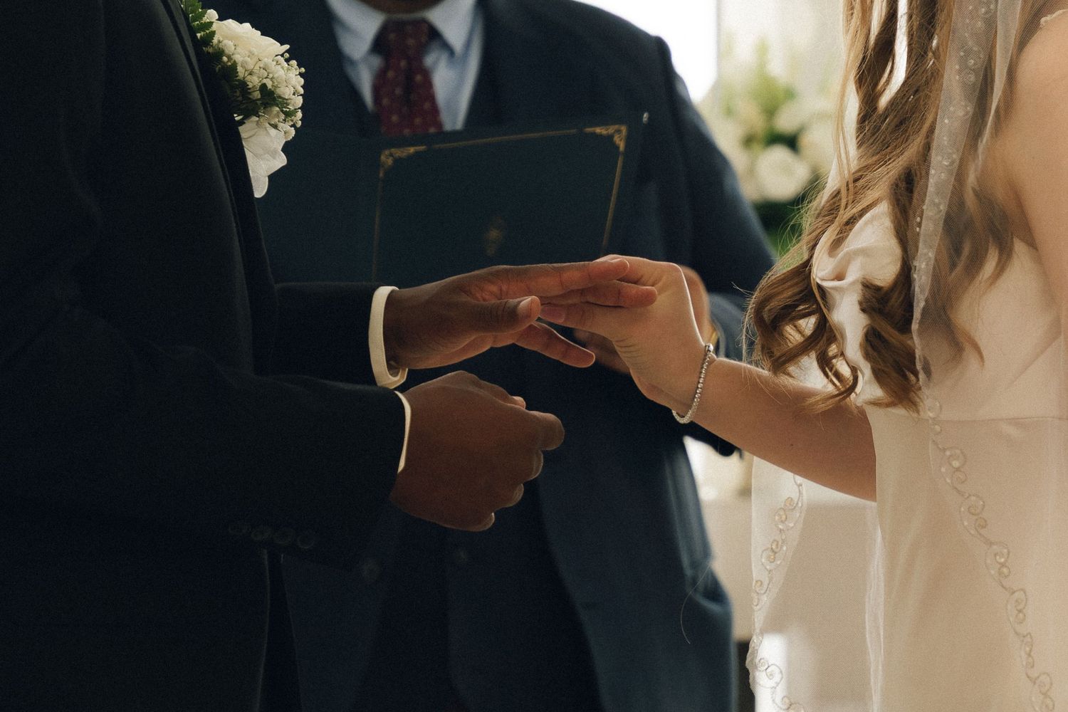 Groom placing a wedding ring on bride's finger during a ceremony, with an officiant holding a book in background.