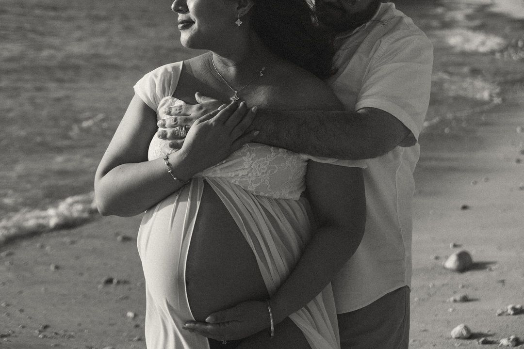 Black and white beach maternity photo of a couple embracing, showcasing pregnant belly by ocean waves.