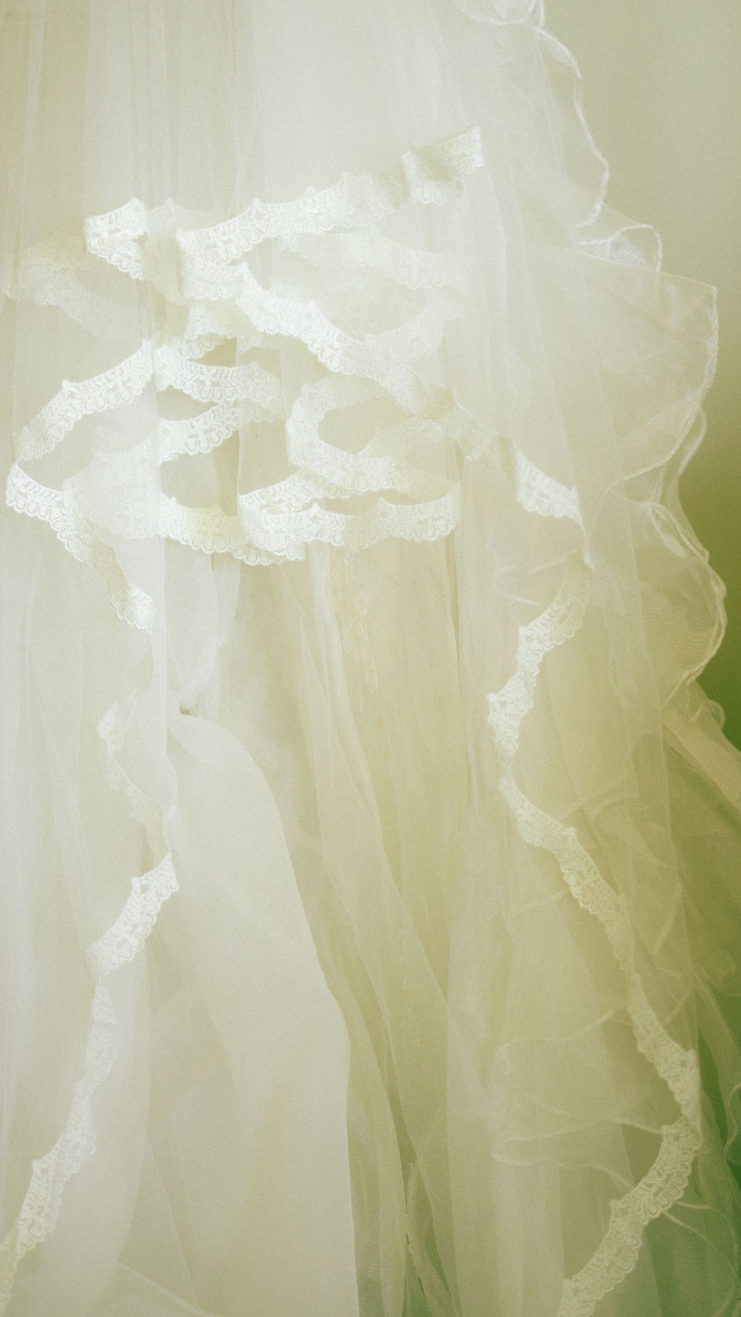 Delicate white tulle wedding veil with ruffled lace trim detail against a soft green background.