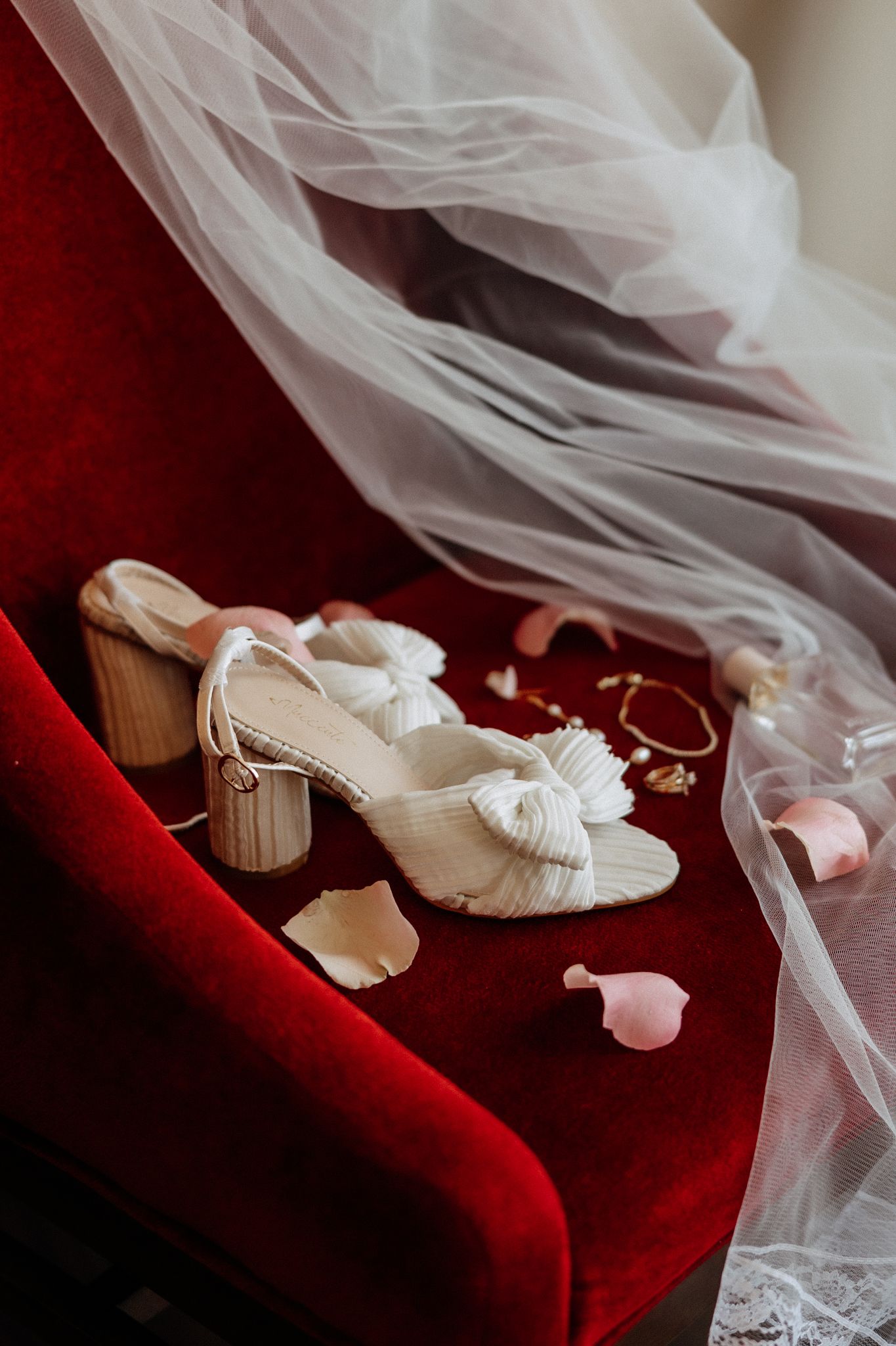 White block heel bridal sandals with bow detail on red velvet chair with veil and petals.