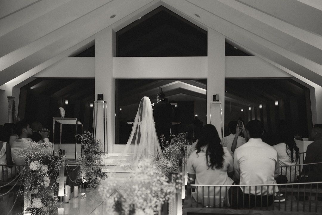 Black and white wedding ceremony photo showing bride at altar with guests seated in elegant chapel venue.