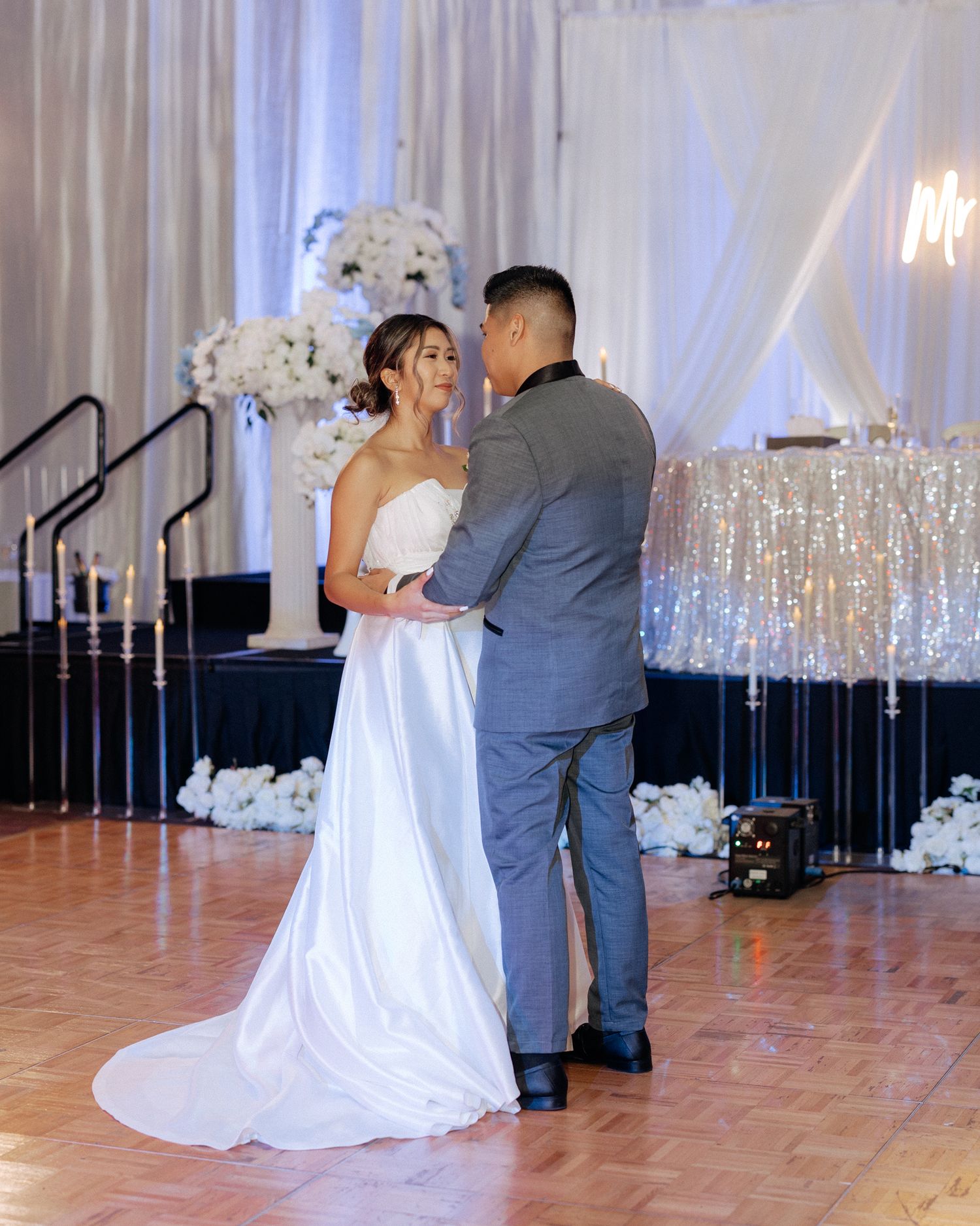 Bride and groom share their first dance on a wooden floor at an elegant wedding reception with white floral decor.