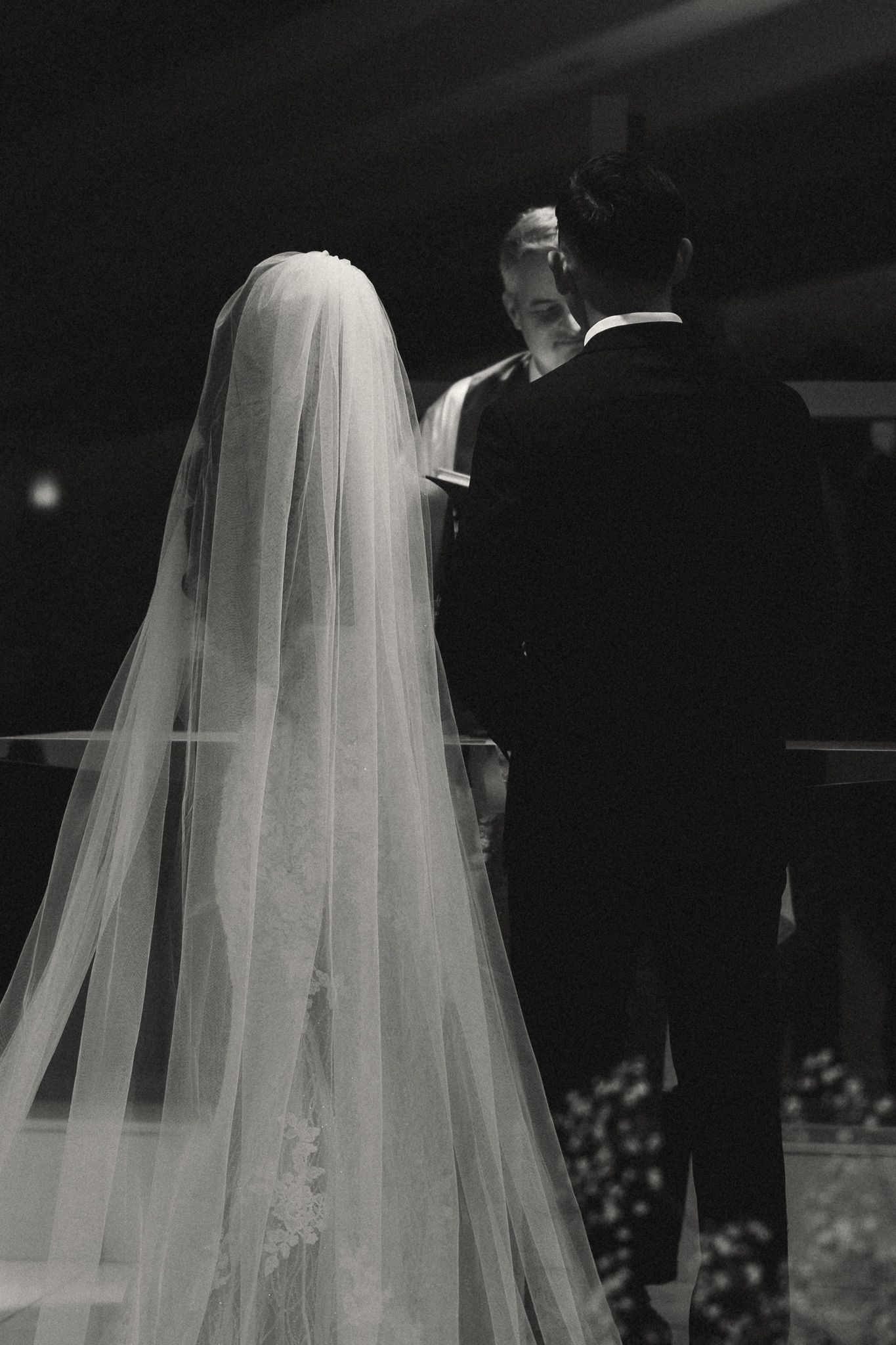 Black and white wedding photo of bride in long veil and groom in tuxedo standing at altar during ceremony.