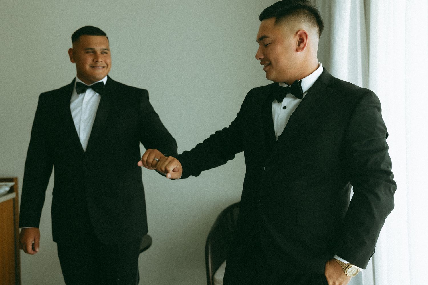Two groomsmen in black tuxedos with bow ties fist bump while getting ready for a wedding.