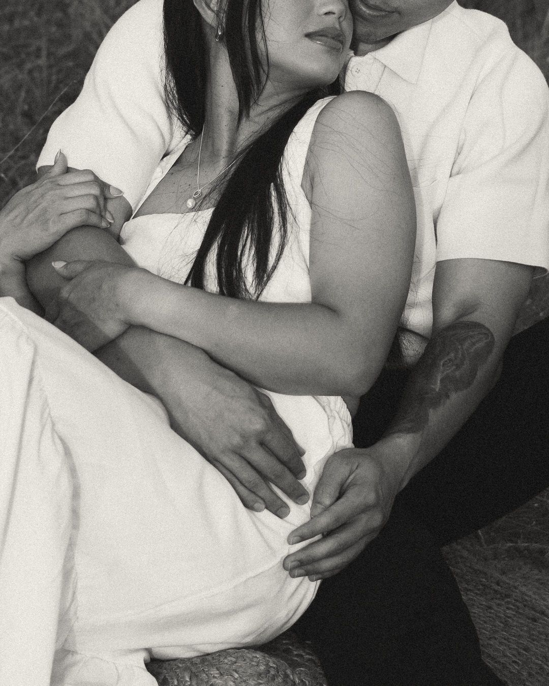 Black and white photo of a couple sitting closely together, man embracing woman in white dress outdoors.