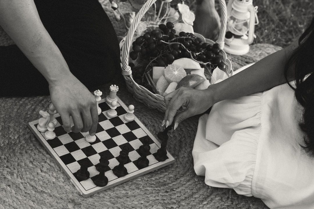Two people playing chess on a checkered board outdoors, with a wicker basket in the background.