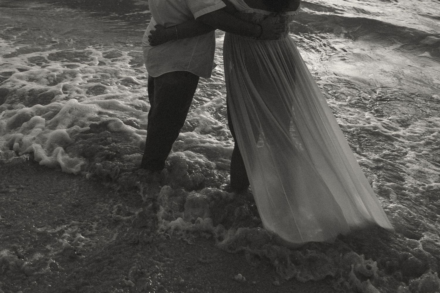 Black and white photo of a couple embracing in ocean waves, woman in flowing white dress, romantic beach scene.