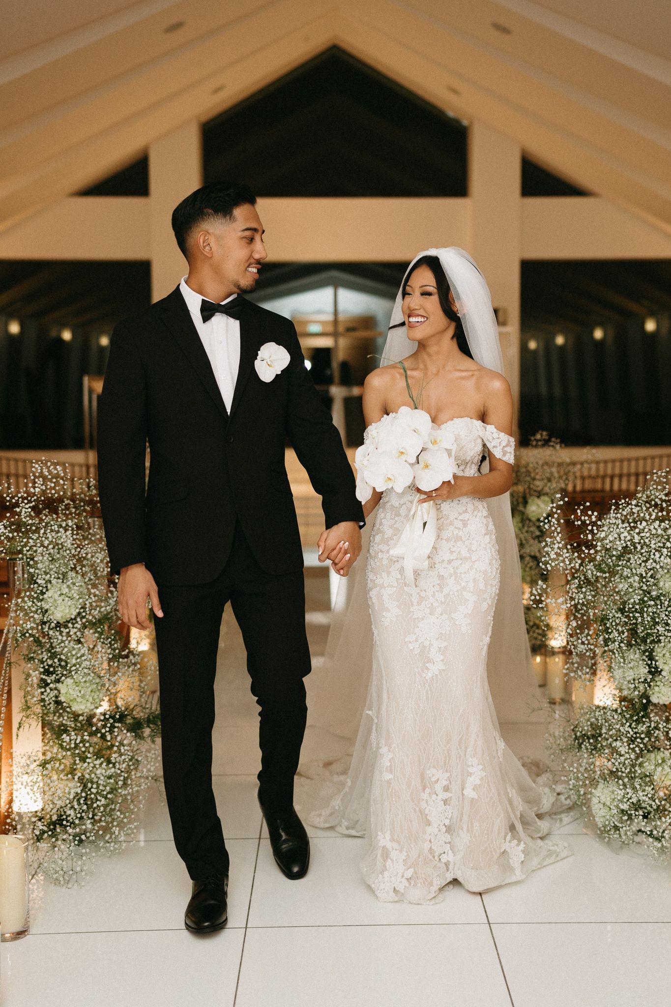 Bride in lace gown and groom in black tuxedo hold hands walking down aisle adorned with candles and baby's breath.
