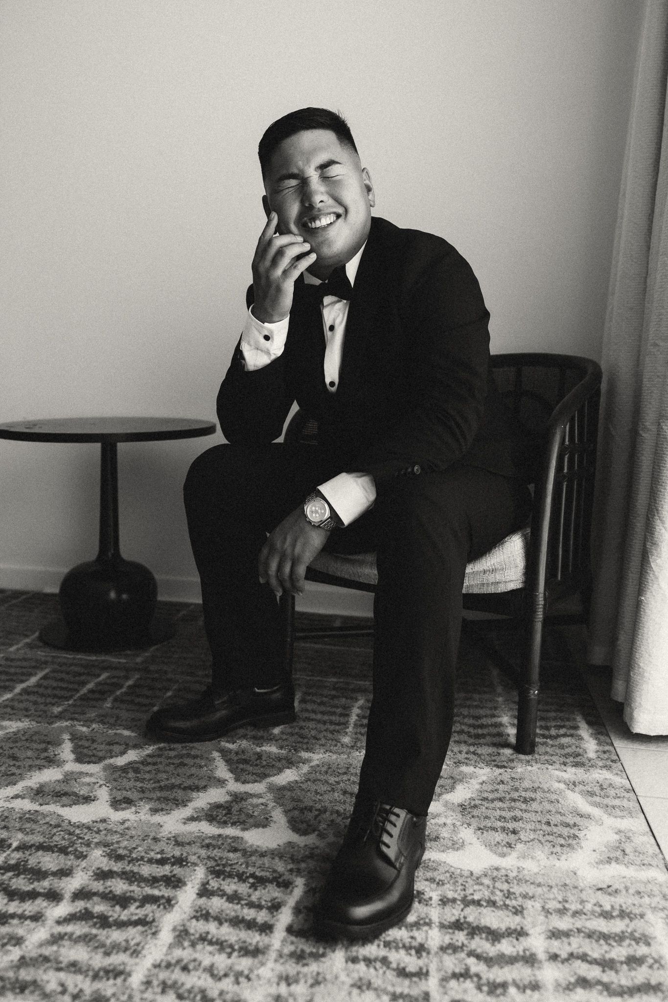Black and white photo of a smiling man in a tuxedo sitting in a chair, laughing candidly.