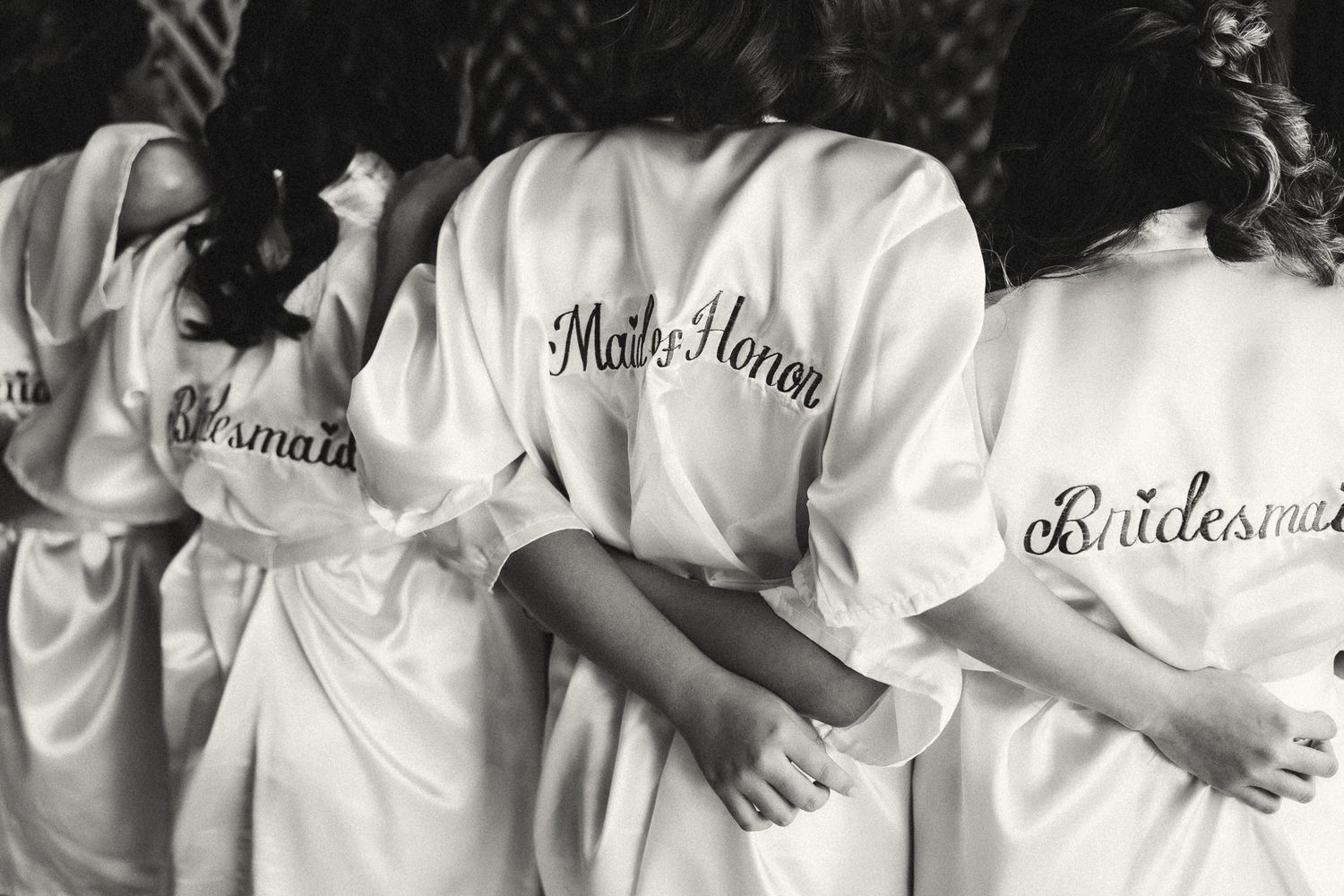 Bridesmaids and maid of honor wearing matching white satin robes with cursive titles on the back.