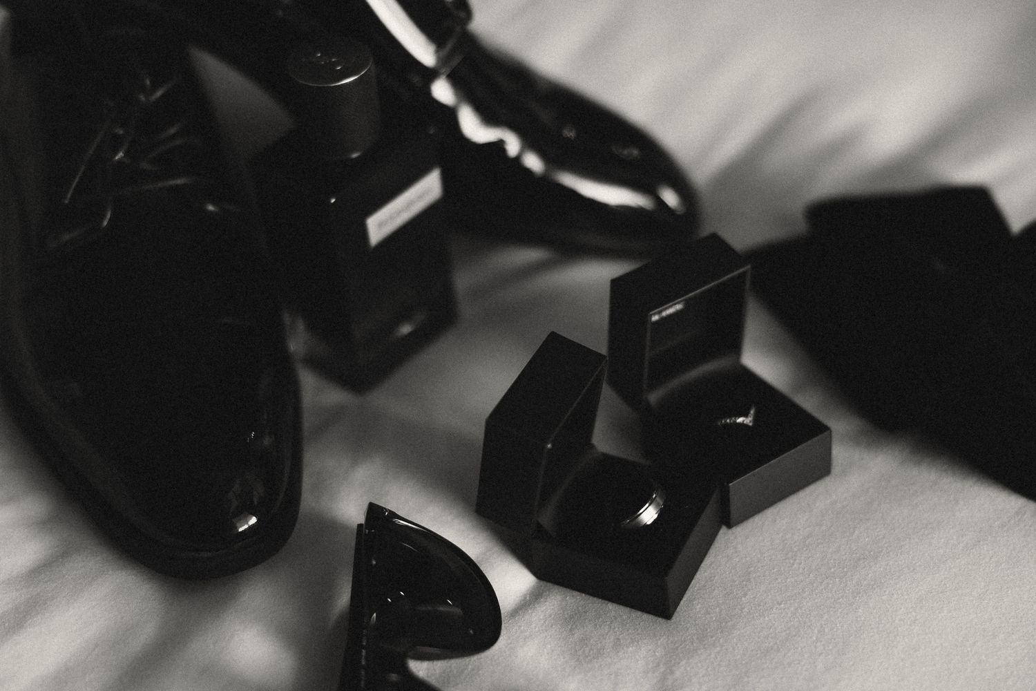 Black and white photo of polished dress shoes, cufflinks, and accessories arranged elegantly on white fabric.