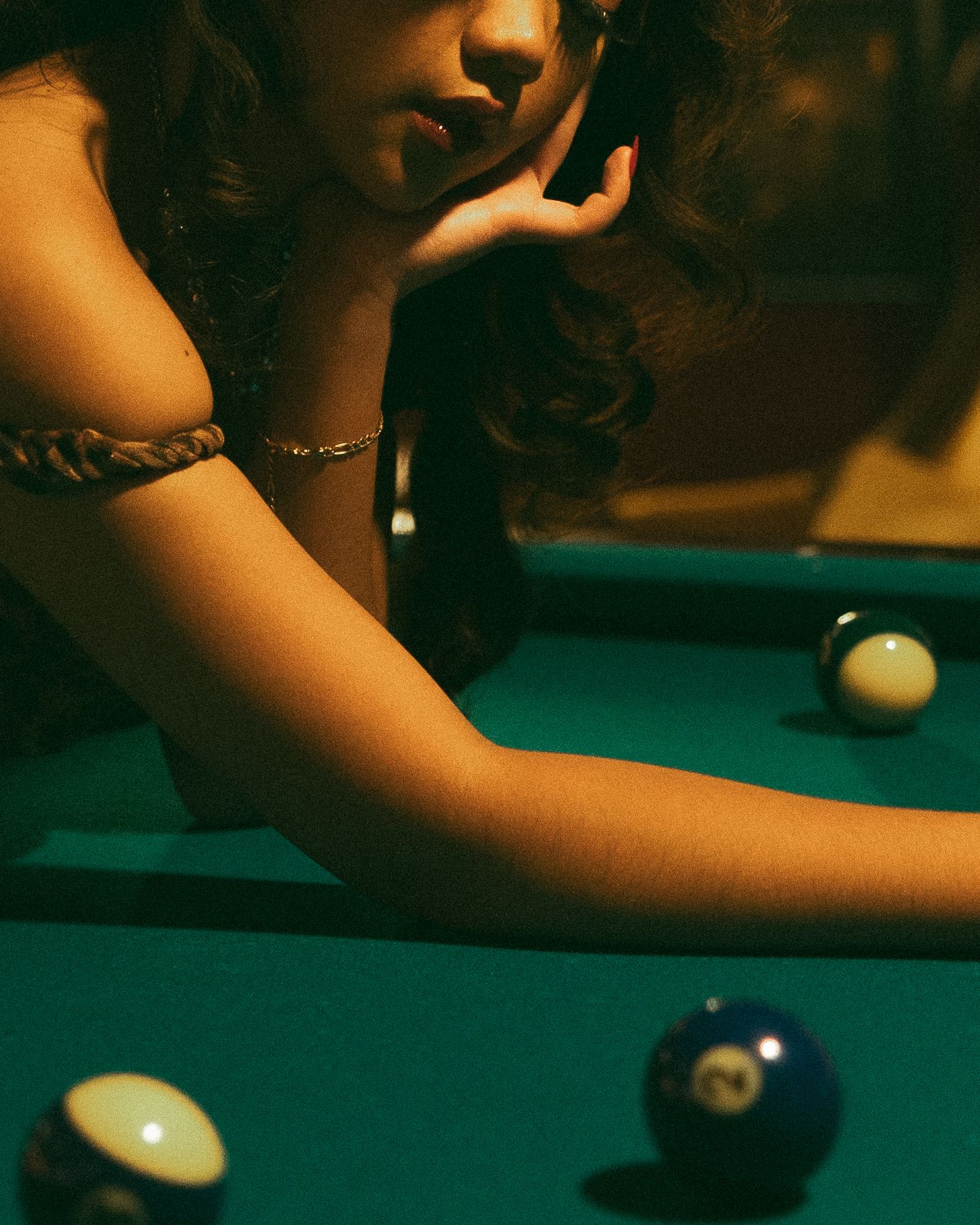 Young woman leaning over a green pool table, resting her chin on her hand, with billiard balls nearby.