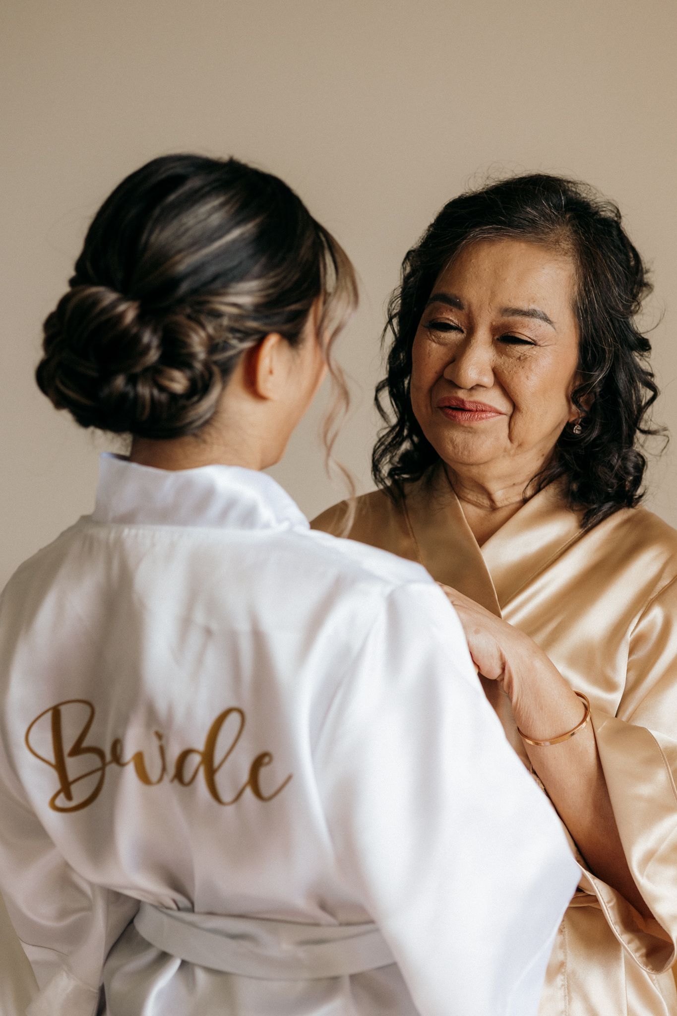 Bride in white satin robe with gold script shares a tender moment with older woman in champagne robe during wedding preparations.