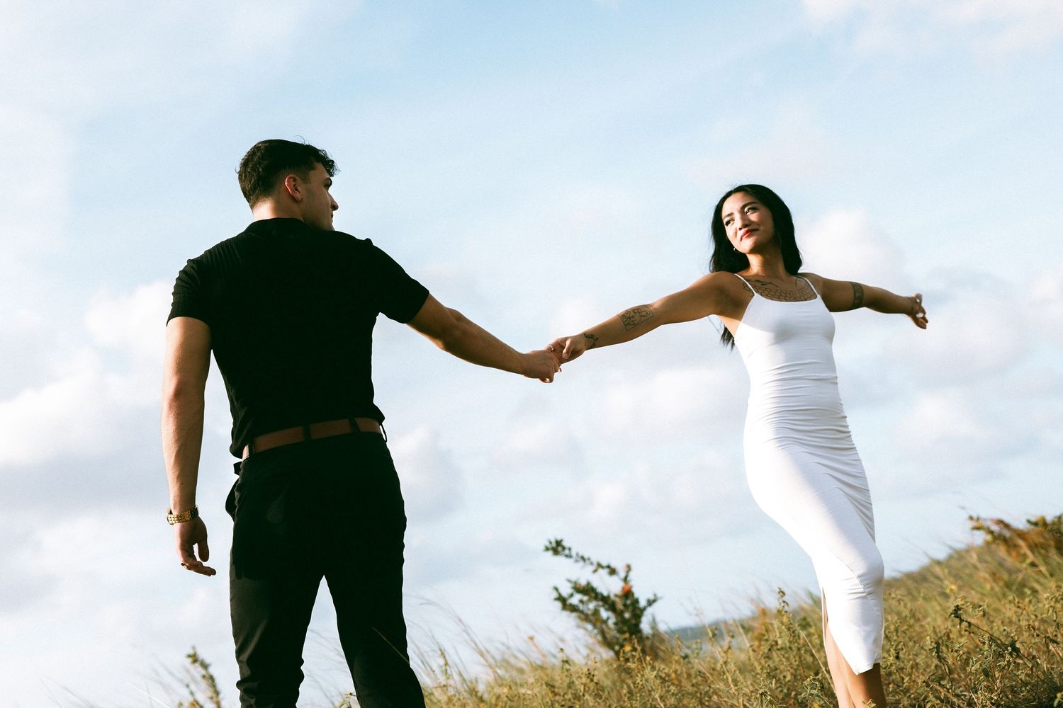 Couple holding hands outdoors, woman in white dress smiling with arms outstretched under a cloudy sky.